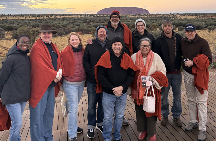 A group of people standing together smiling. Uluru is in the background. 